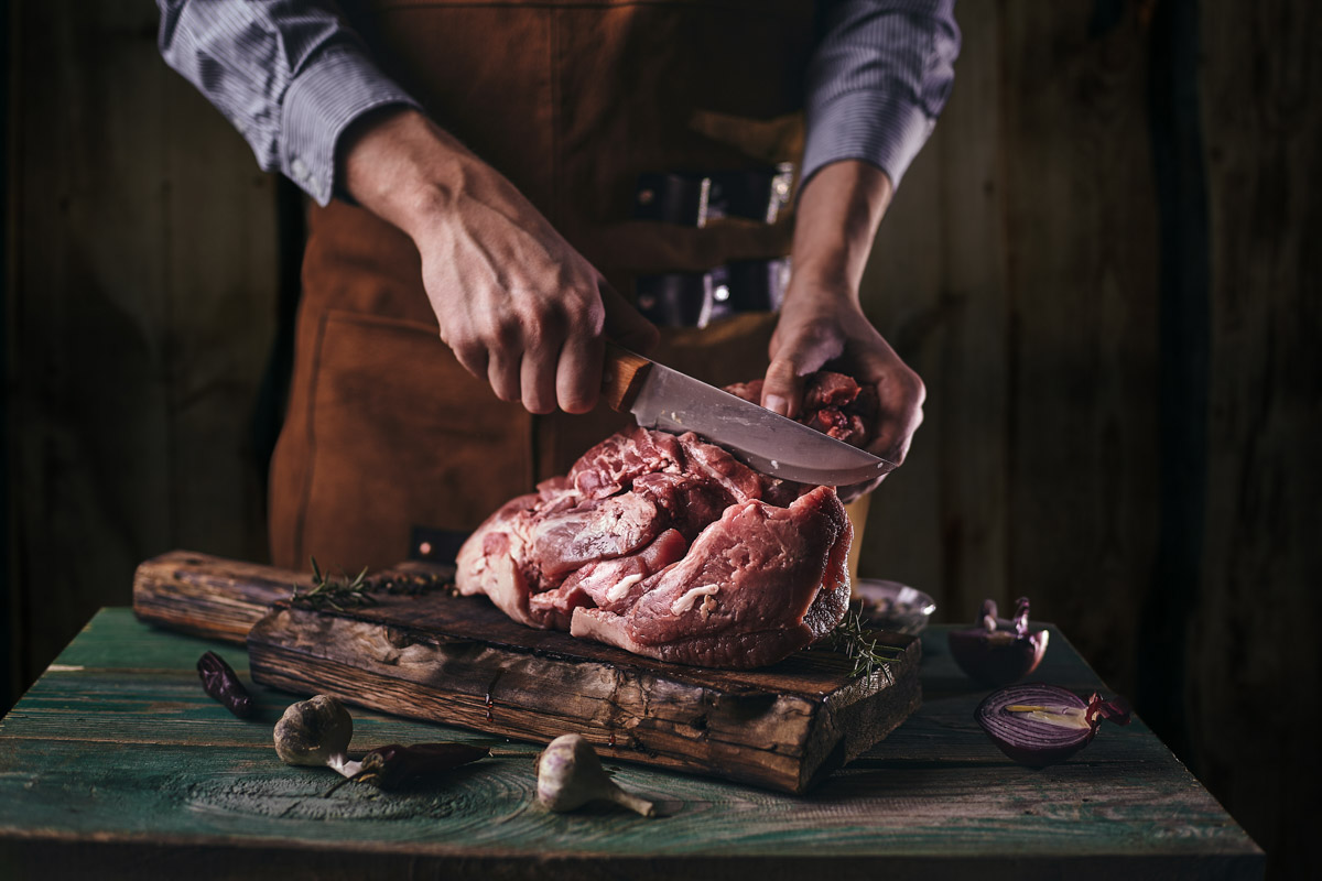 A butcher in a leather apron cuts a large piece of meat on a wooden board.